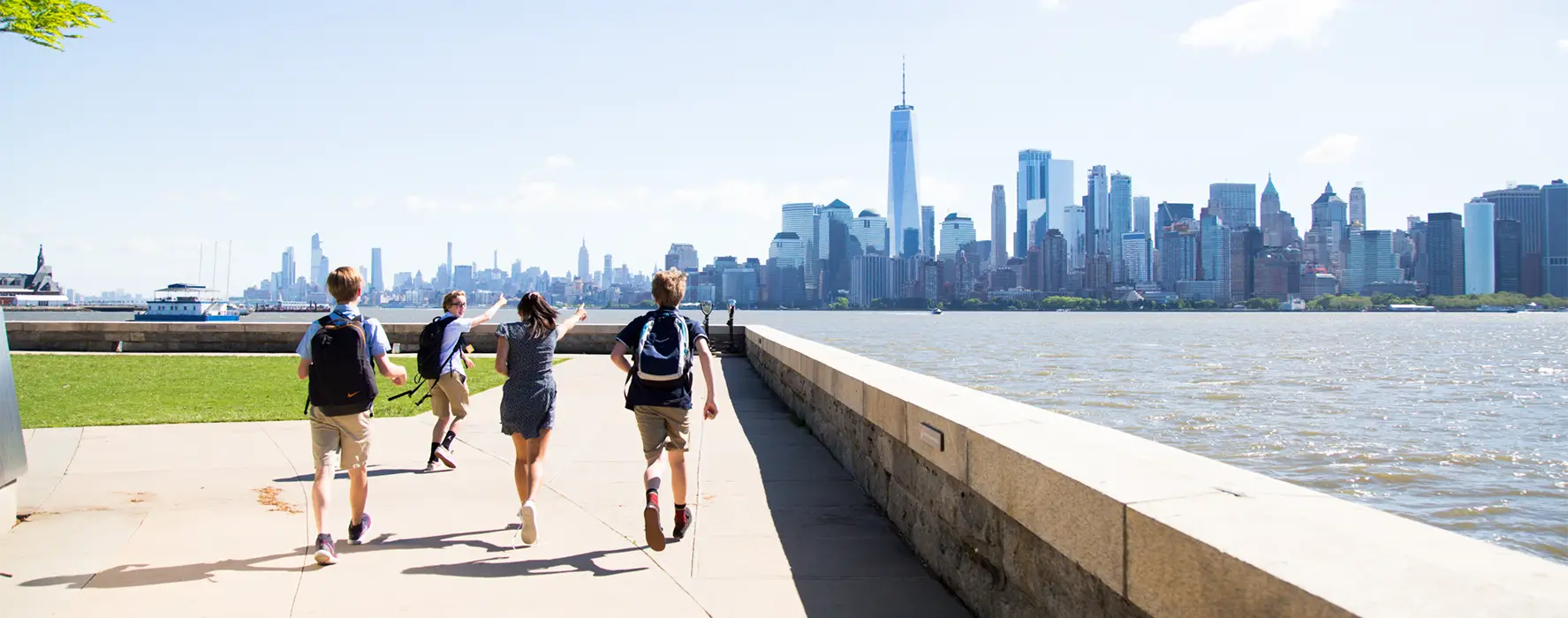 A group of 4 students running down a sidewalk pointing at the NYC skyscrapers across the water.