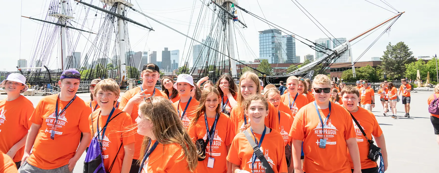 A group of students in orange shirts standing in front of ships with their sails lowered, with the Boston skyline in the background.