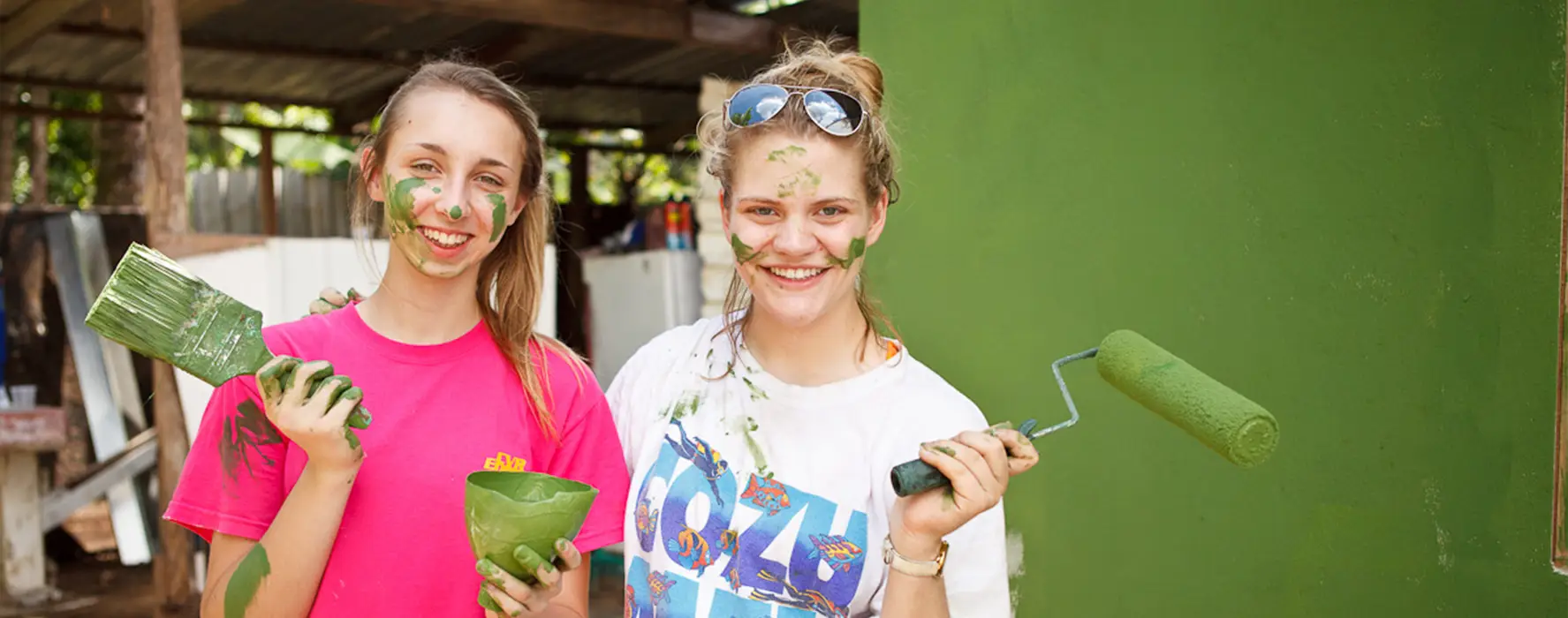 Two students painting a wall, with green paint splashes on their faces.