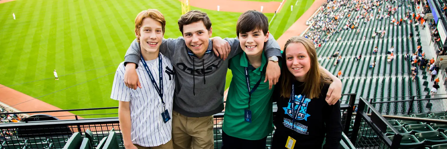 Four students posing for a photo in the stands of a baseball stadium with a game happening in the background.